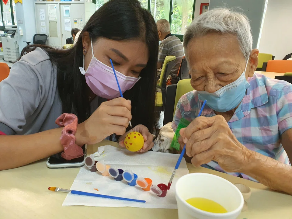 A PSA Health@Home volunteer engaging a senior from St Luke’s ElderCare Telok Blangah Centre in painting Easter eggs.
