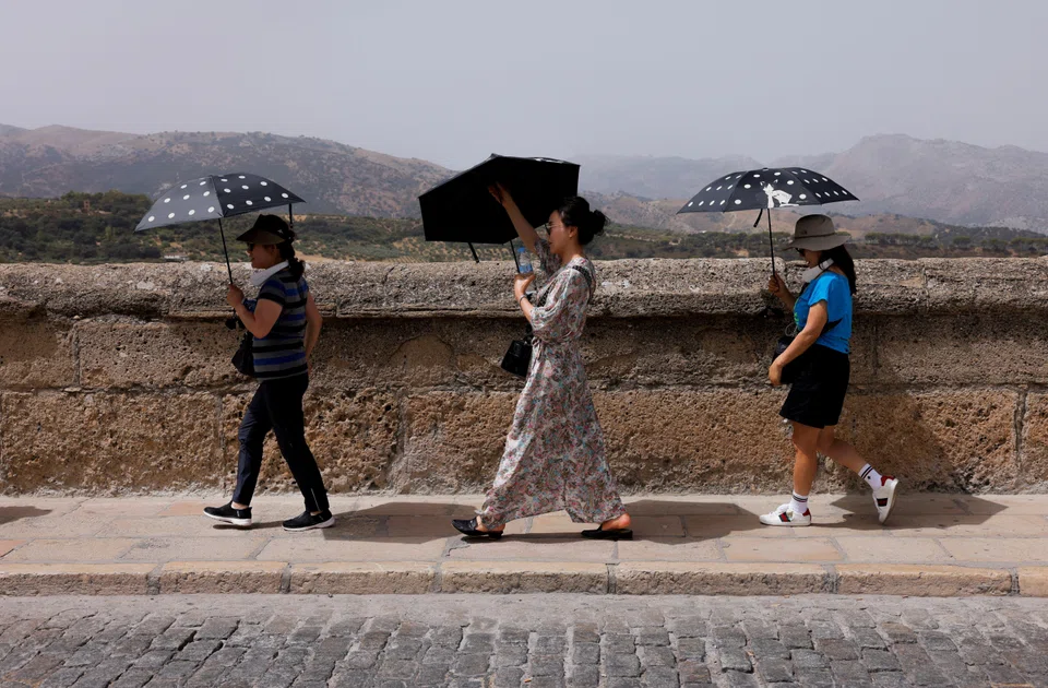 Tourists shield themselves with umbrellas, under a sky which is hazy due to suspended sand particles from the Sahara desert, a weather phenomenon known locally as 'Calima', during Spain's third heatwave of the summer, in Ronda, Spain, Aug 9, 2023. 