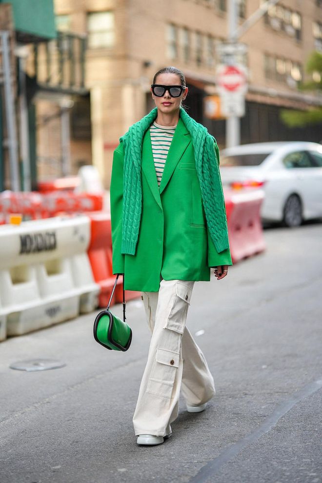 NEW YORK, NEW YORK - SEPTEMBER 10: A guest wears black sunglasses, a beige and green striped print pattern t-shirt, a green oversized blazer jacket, a green braided wool pullover, white latte denim cargo pants, a green shiny leather and black borders handbag, white leather pointed pumps heels shoes , outside Tibi , during New York Fashion Week, on September 10, 2022 in New York City. (Photo by Edward Berthelot/Getty Images)