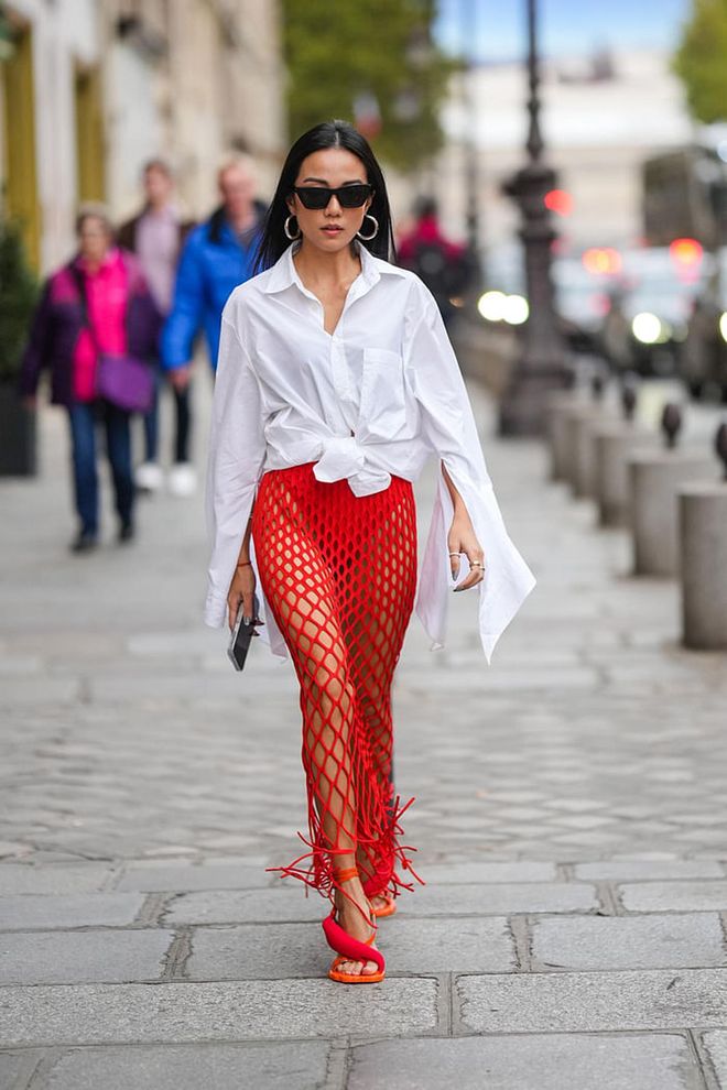 PARIS, FRANCE - SEPTEMBER 30: Yoyo Cao wears black sunglasses, silver earrings, a white oversized shirt, a red mesh / fishnet long fringed skirt, silver rings, orange shiny leather strappy / red puffy strap heels mules , outside Alessandra Rich, during Paris Fashion Week - Womenswear Spring/Summer 2023, on September 30, 2022 in Paris, France. (Photo by Edward Berthelot/Getty Images)