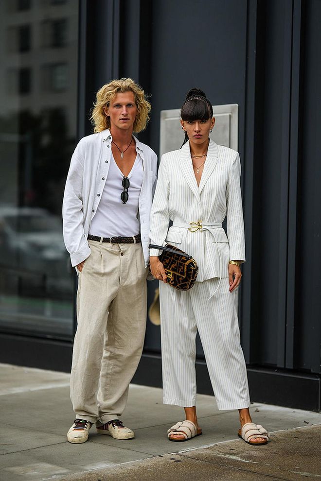 NEW YORK, NEW YORK - SEPTEMBER 11: Luca Froehlingsdorf (L) wears a black with silver pendant necklace, a white V-neck tank-top, a white velvet shirt, black sunglasses, a brown and black braided leather belt, beige linen suit pants, beige with multicolored striped print pattern sneakers, Katya Tolstova (R) wears silver earrings, a silver and gold large chain necklace, a gold chain pendant necklace, a white with black small striped print pattern long blazer jacket, matching white with black small striped print pattern large pants, a white shiny leather with gold large buckle belt, a gold large bracelet, a brown and black FF monogram print pattern fluffy handbag from Fendi, white sheep fluffy strappy sandals , during New York Fashion Week, on September 11, 2022 in New York City. (Photo by Edward Berthelot/Getty Images)
