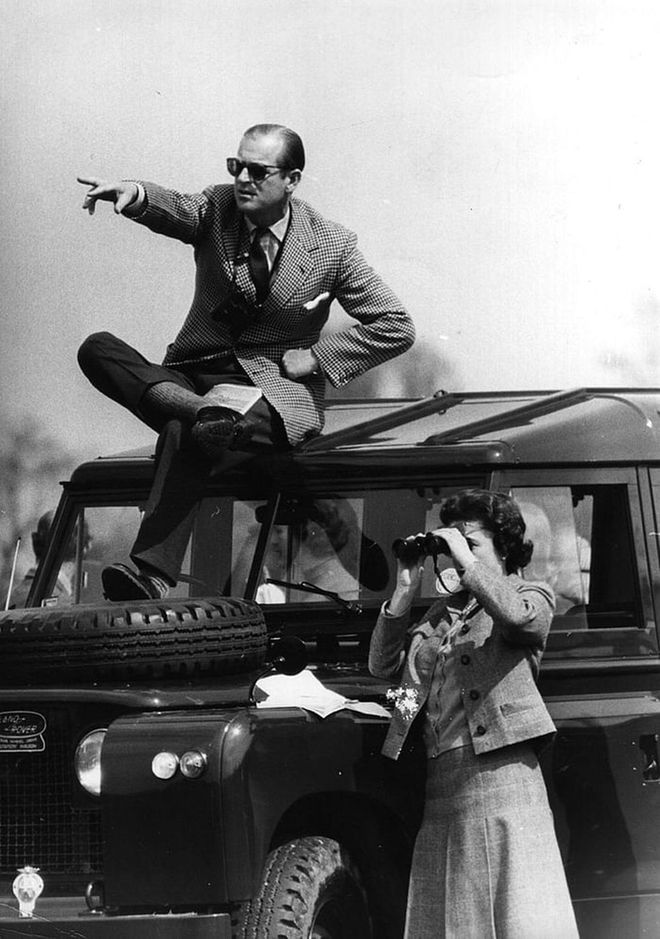 Prince Philip on top of a Land Rover pointing out competitors at the Badminton Horse Trials to Queen Elizabeth II as she watches through binoculars.