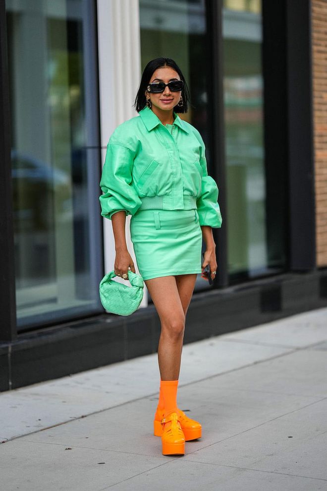 NEW YORK, NEW YORK - SEPTEMBER 10: A guest wears black sunglasses from Gucci, gold large logo earrings from Valentino, a green zipper / puffy sleeves bomber coat, a matching green short skirt, a matching green shiny braided leather Joddie handbag from Bottega Veneta, silver and gold earrings, neon orange socks from Loewe, neon orange shiny leather strappy plastic meduse shoes from Loewe, outside Tibi , during New York Fashion Week, on September 10, 2022 in New York City. (Photo by Edward Berthelot/Getty Images)