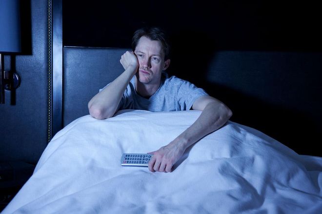 Photo of a man sitting in his bed, watching late night television with remote control in hand.