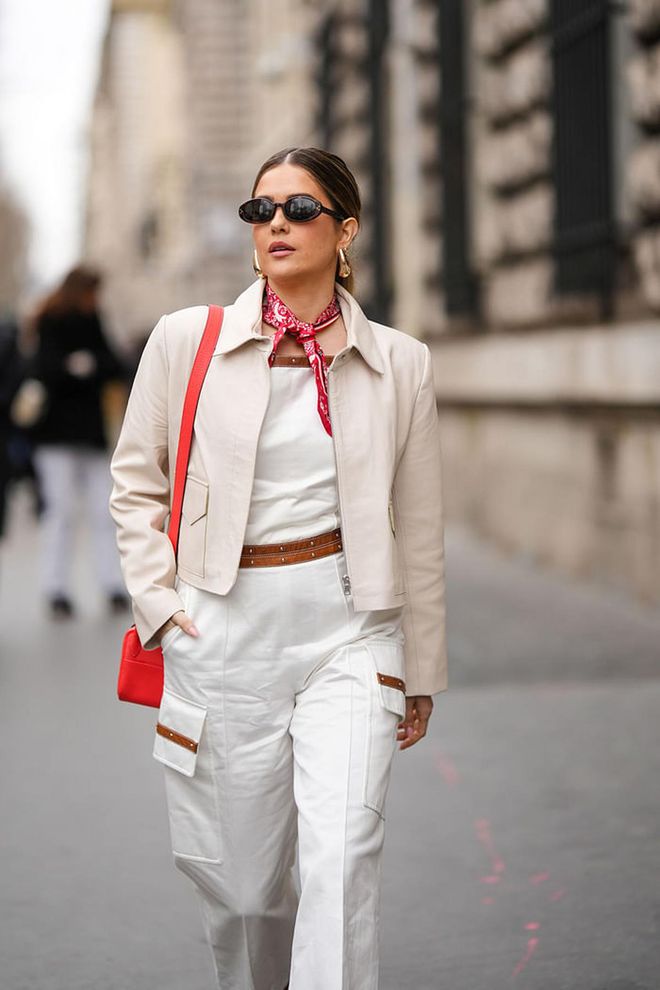 PARIS, FRANCE - MARCH 04: Paola Alberdi wears black sunglasses, a white and red print pattern silk scarf, a white shoulder-off / denim wide legs pants with a camel belt / cargo jumpsuit, a white matte leather zipper jacket, a neon red shiny leather shoulder bag, outside Hermes, during Paris Fashion Week - Womenswear Fall Winter 2023 2024, on March 04, 2023 in Paris, France. (Photo by Edward Berthelot/Getty Images)