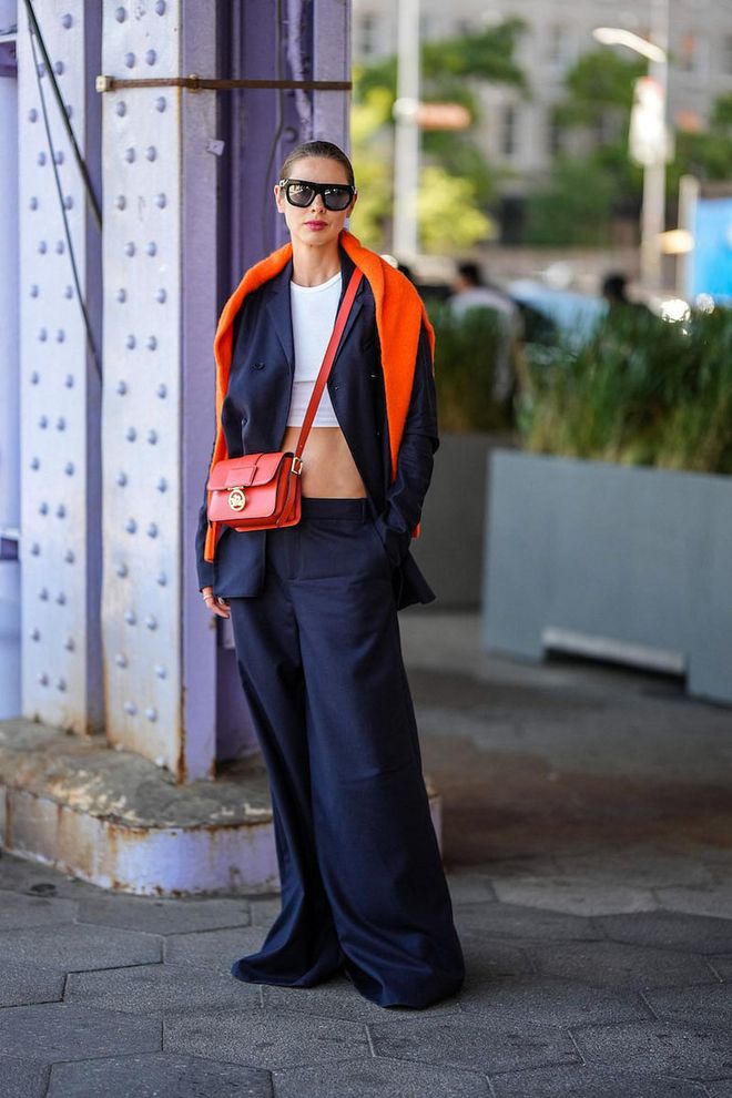 NEW YORK, NEW YORK - SEPTEMBER 10: A guest wears black sunglasses, a neon orange wool pullover, a white cropped t-shirt, a navy blue blazer jacket, matching navy blue wide legs pants, a red shiny leather crossbody bag from Longchamp, outside Jason Wu, during New York Fashion Week, on September 10, 2022 in New York City. (Photo by Edward Berthelot/Getty Images)