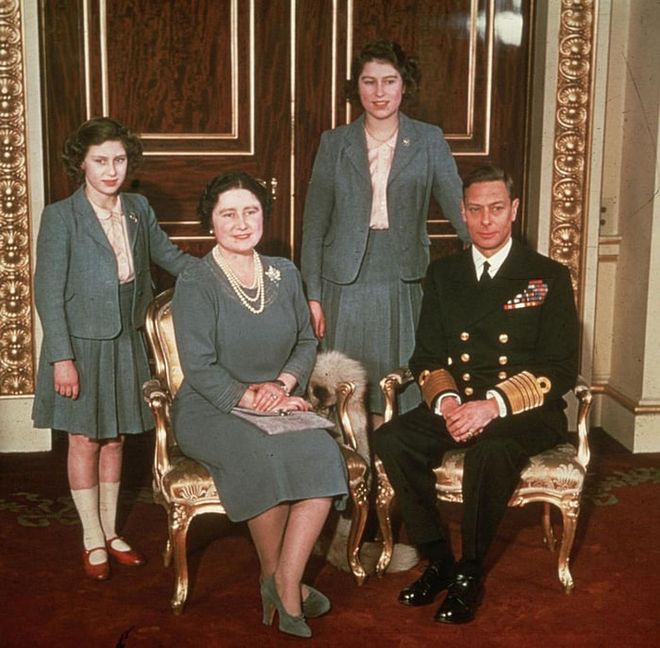 Princess Margaret, Queen Elizabeth the Queen Mother, Princess Elizabeth, and King George VI pose for a family portrait at Buckingham Palace. The royal daughters match in gray blazers, pleated skirts, and peach button-down shirts.