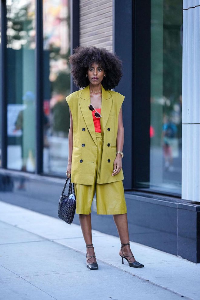 NEW YORK, NEW YORK - SEPTEMBER 10: A guest wears silver earrings, beige sunglasses, a red shoulder-off top, a green sleeveless / buttoned oversized jacket, matching green shiny leather large shorts, a black pearls handbag, black shiny leather laces ankle / block heels ballerinas, a silver and gold watch, outside Tibi , during New York Fashion Week, on September 10, 2022 in New York City. (Photo by Edward Berthelot/Getty Images)