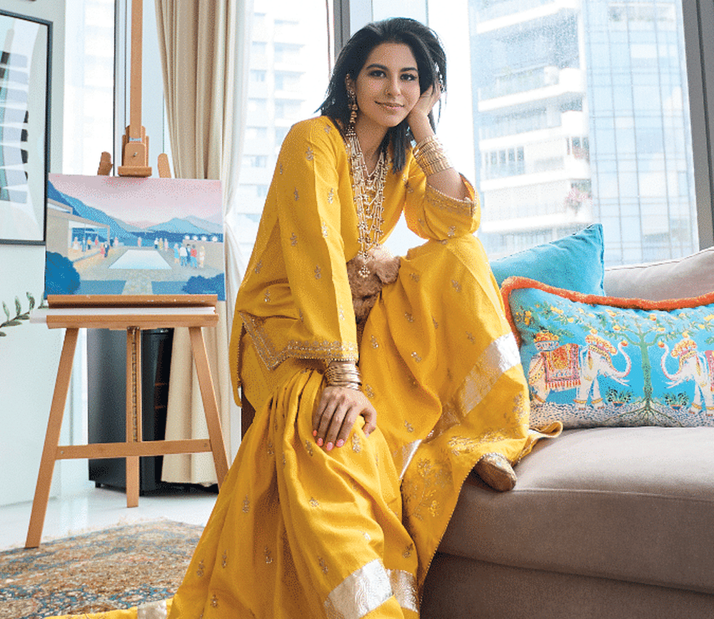 Perched on her living room sofa in front of a painting by her grandfather, Kausar is dressed in traditional wide-legged gharara trousers paired with a kurta, a sat lara necklace and earrings gifted by her in-laws on her first visit to India, together with SunMoonRain bangles, and embroidered traditional shoes from Pakistan