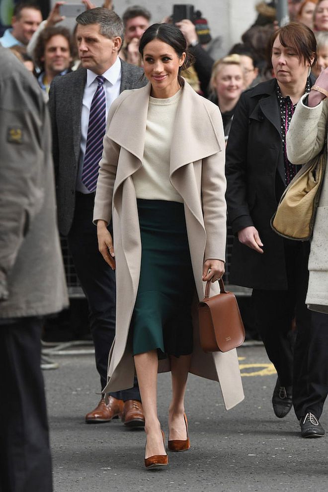 Meghan visited Northern Ireland in a Mackage Mai wool belted coat, left undone. Underneath she chose an ivory cashmere sweater by personal friend, Victoria Beckham and a forest green pencil skirt by Greta Constantine. Her brown Charlotte Elizabeth bag matches her Jimmy Choo pumps perfectly! She kept it casual and controversial by showing up with a messy bun too, as most royals would wear hair nets. 
