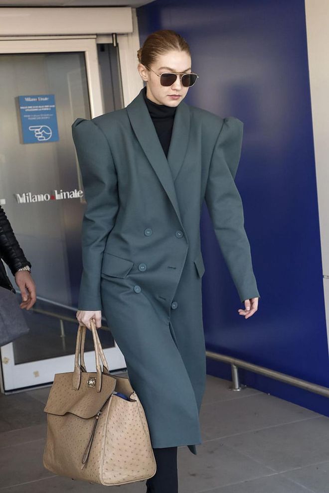 Hadid touched down in Milan wearing a teal-gray trench coat with bold shoulders, black turtleneck, black shades, and carried an ostrich Hermès bag.