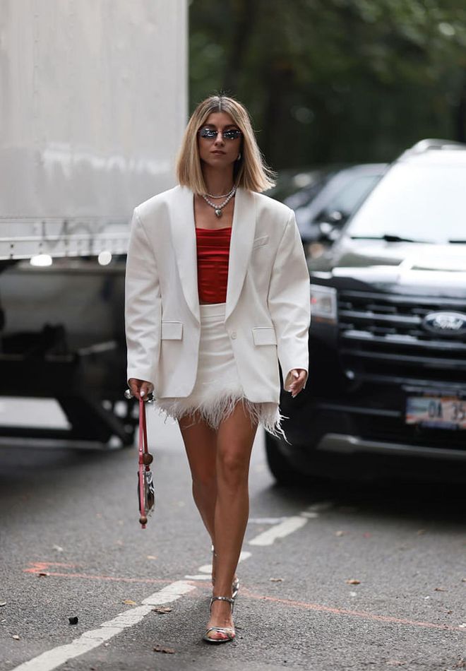 NEW YORK, NEW YORK - SEPTEMBER 08: A Fashion Week Guest is seen wearing narrow sunglasses with a red frame, pearl necklaces, a white oversized blazer, underneath a satin corset top in red, a white feathered mini skirt, a huge silver ring, a shiny silver handbag from Jacquemus and silver strap heels outside before the Christian Siriano Show on September 08, 2023 in New York City. (Photo by Jeremy Moeller/Getty Images)