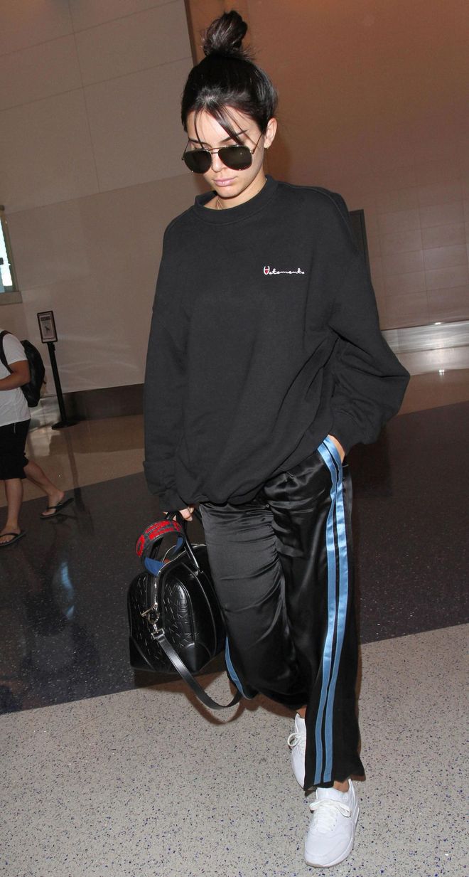In a Vetements sweatshirt, black satin track pants, white sneakers, a black leather satchel and aviator sunglasses in LAX.