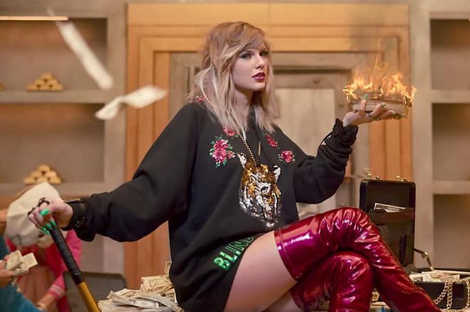 Very cold from not wearing pants, bank-robbing Taylor sets fire to a pile of cash to keep warm, while showing off her bedhead-y waves and glitter-flecked burgundy lips.